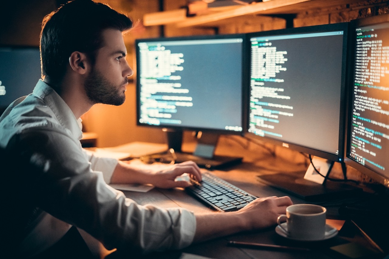Focused young developer coding late in office writing script shown on computer monitors, serious handsome coder programmer hacker programming developing software applications working alone at night